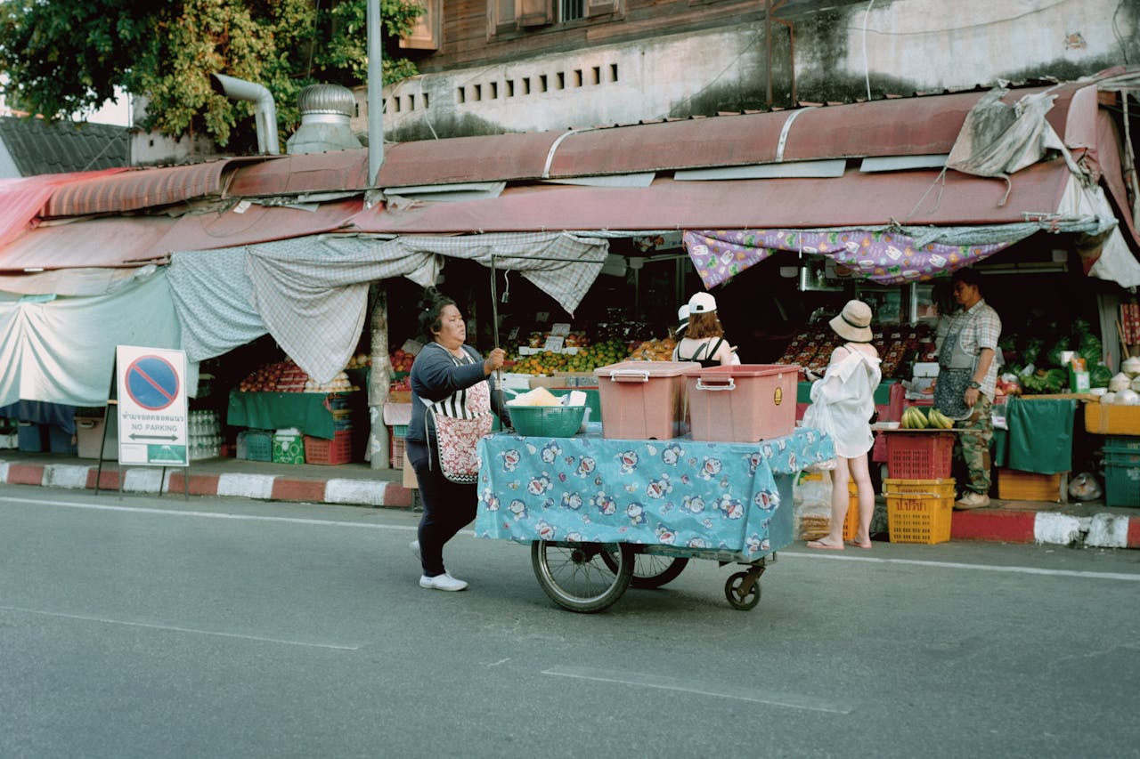 A bustling urban street market with a vendor pushing a cart, surrounded by market stalls and pedestrians.